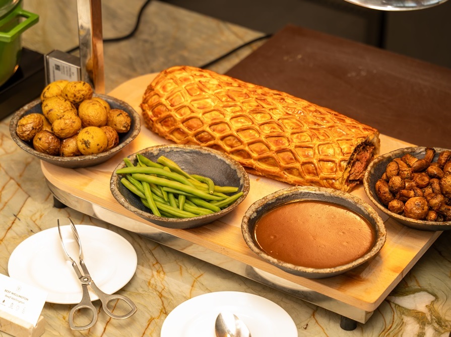 Golden puff pastry meat pie on a wooden board with bowls of roasted potatoes, green beans, gravy, and roasted mushrooms.