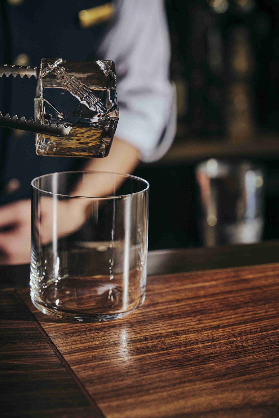 Bartender places a large ice cube into a lowball glass on a wooden bar.