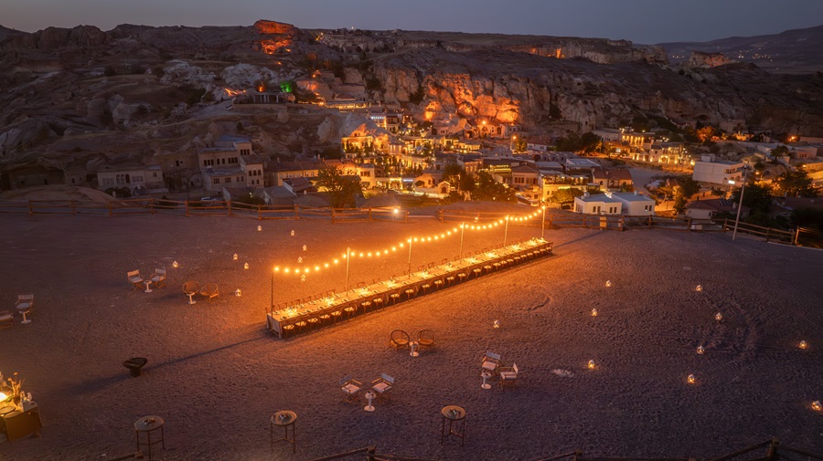 Evening view of a beachside dining area with a long row of tables under string lights, overlooking a hillside town illuminated in warm lights.”] ,
