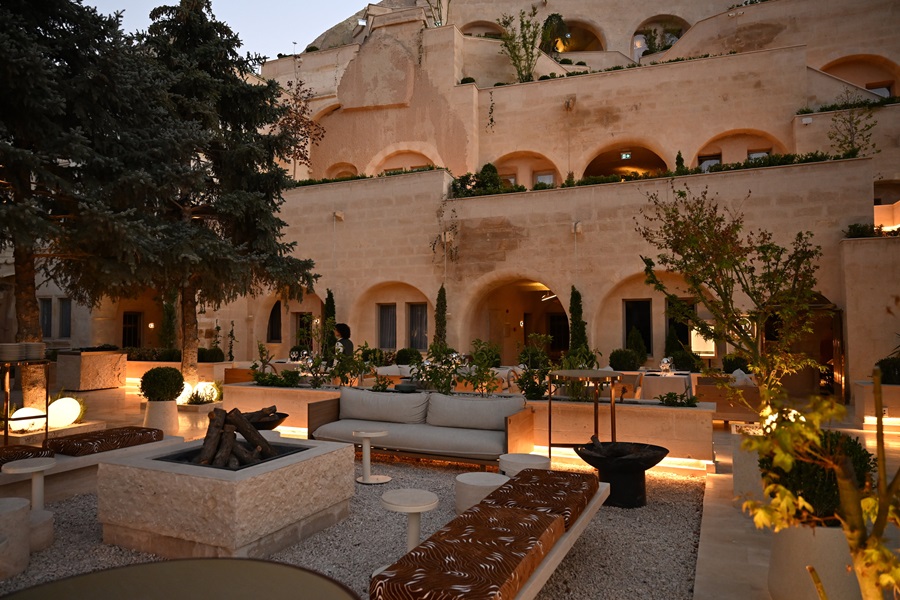 Beige stone hotel courtyard at dusk with arches, trees, and a central square fire pit surrounded by seating.