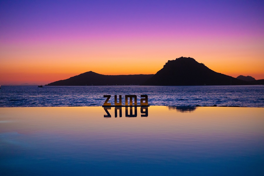Sunset over the ocean with silhouetted hills, calm water reflecting the vibrant sky and the word 'ZUMA' in the foreground.