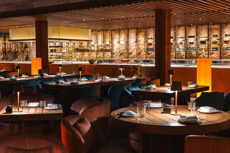 Warm, contemporary restaurant interior with round wooden tables, cushioned chairs, and ambient orange lighting; open kitchen in the background
