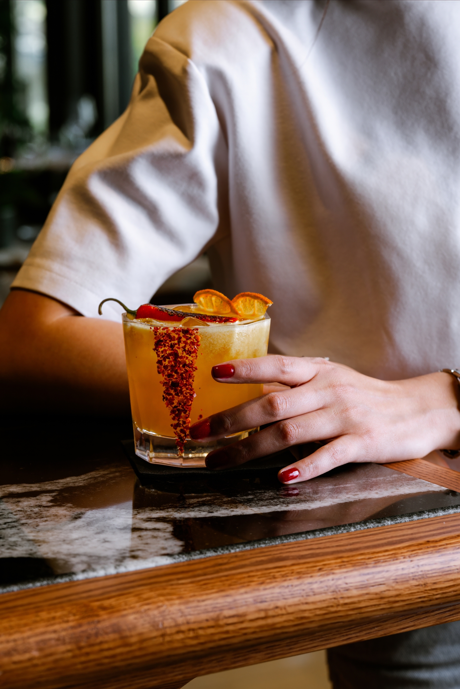 Person in a light sweater holds a yellow cocktail with orange slices and a chili-salt rim on a bar.