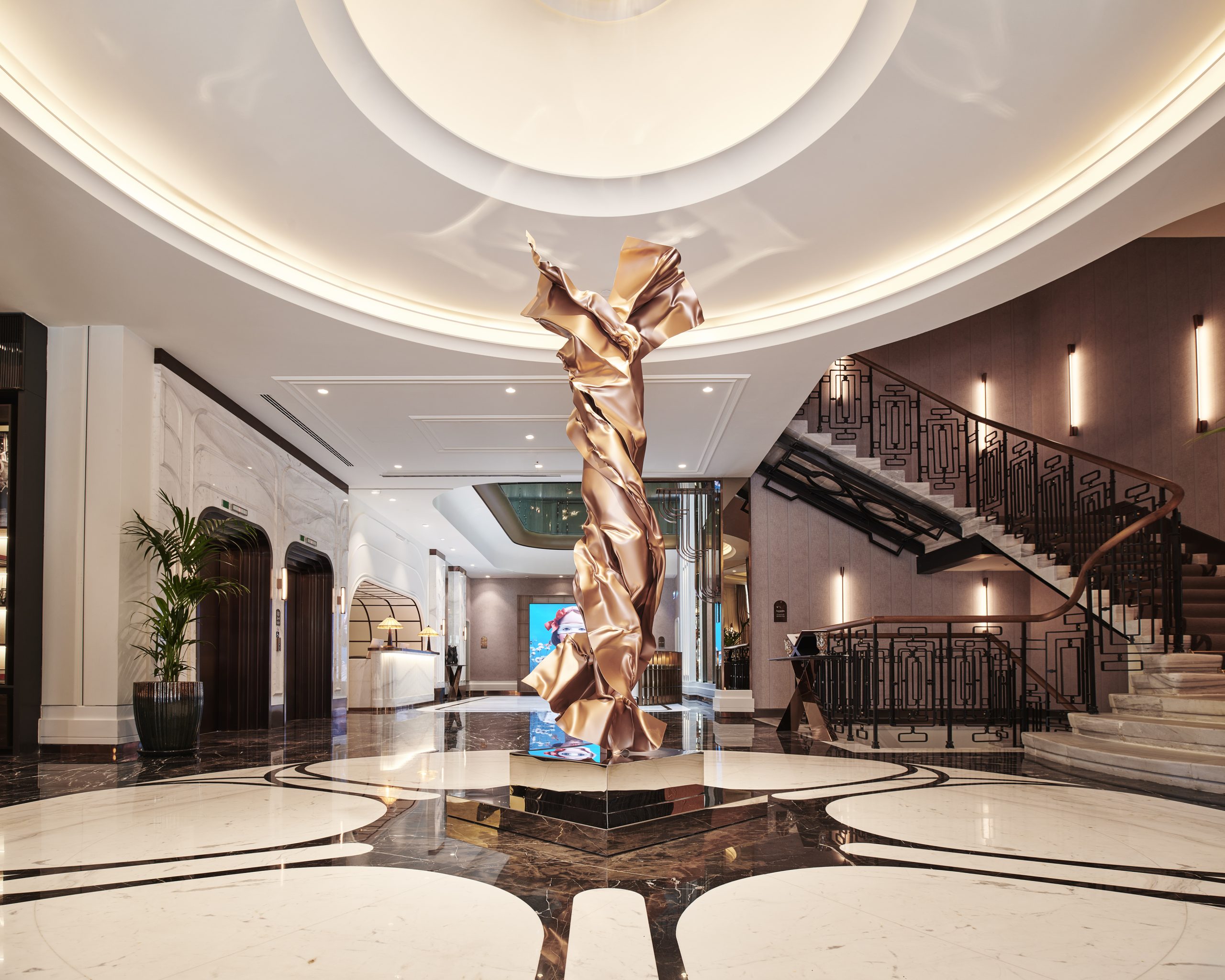 Les Temps_Lobby 13 | HotelRestaurant Haber Dergisi Luxurious hotel lobby with a twisting metallic sculpture in gold at center, circular ceiling rings above, and a grand staircase to the right.