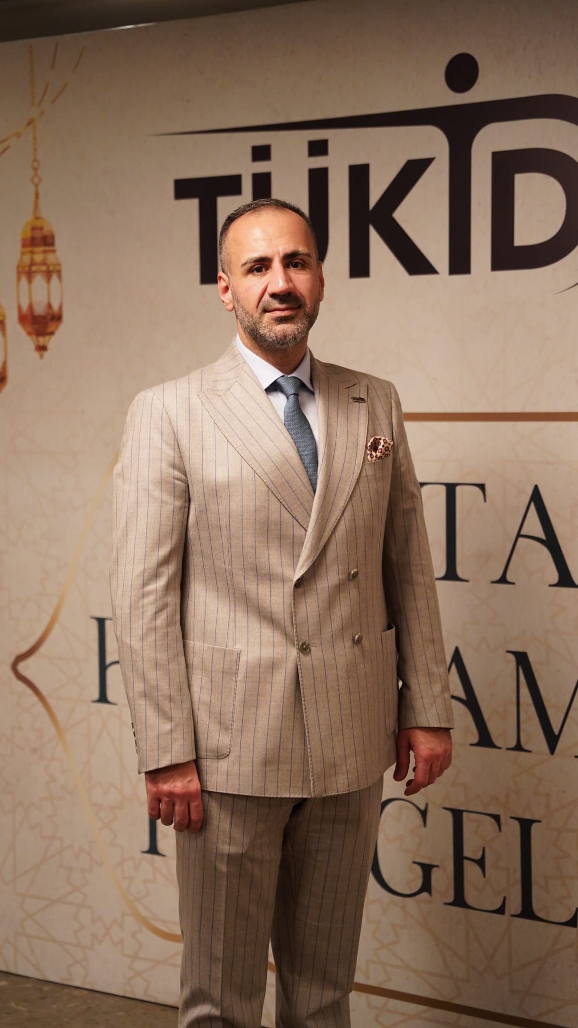 Man in a beige pinstripe suit posing at an event backdrop with large black text behind him, blue tie and pocket square.