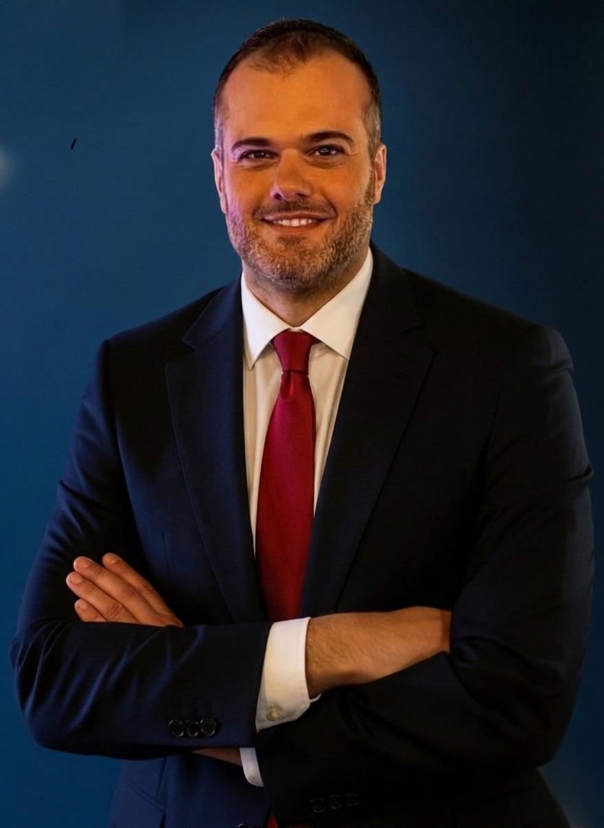 Professional headshot of a man in a dark suit and red tie, arms crossed, against a blue background, smiling confidently.