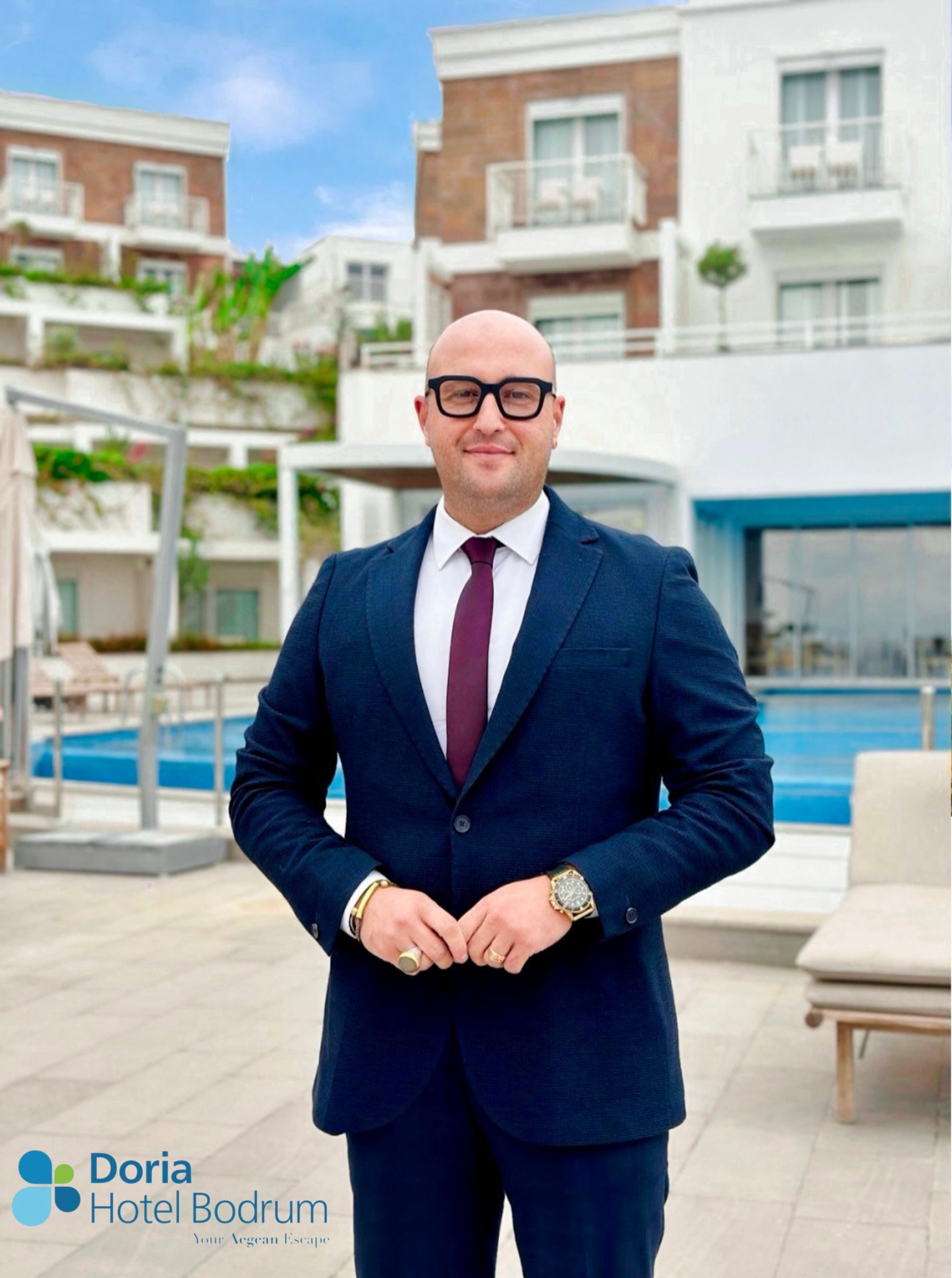Man in a navy suit and maroon tie posing outdoors by a pool with modern white buildings in the background; logo for Doria Hotel Bodrum appears in the corner.