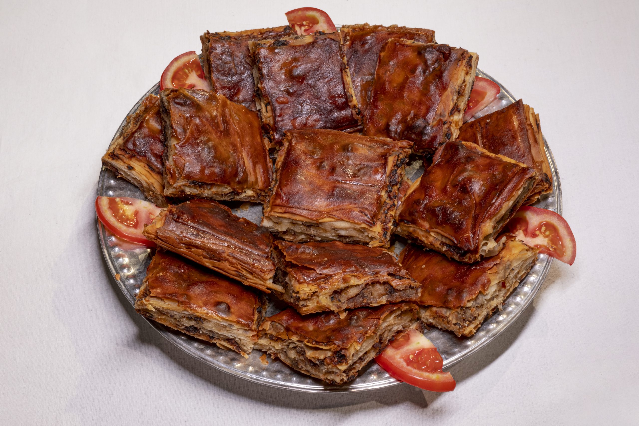 Stack of honey-soaked baklava squares on a metal platter with tomato wedges around them