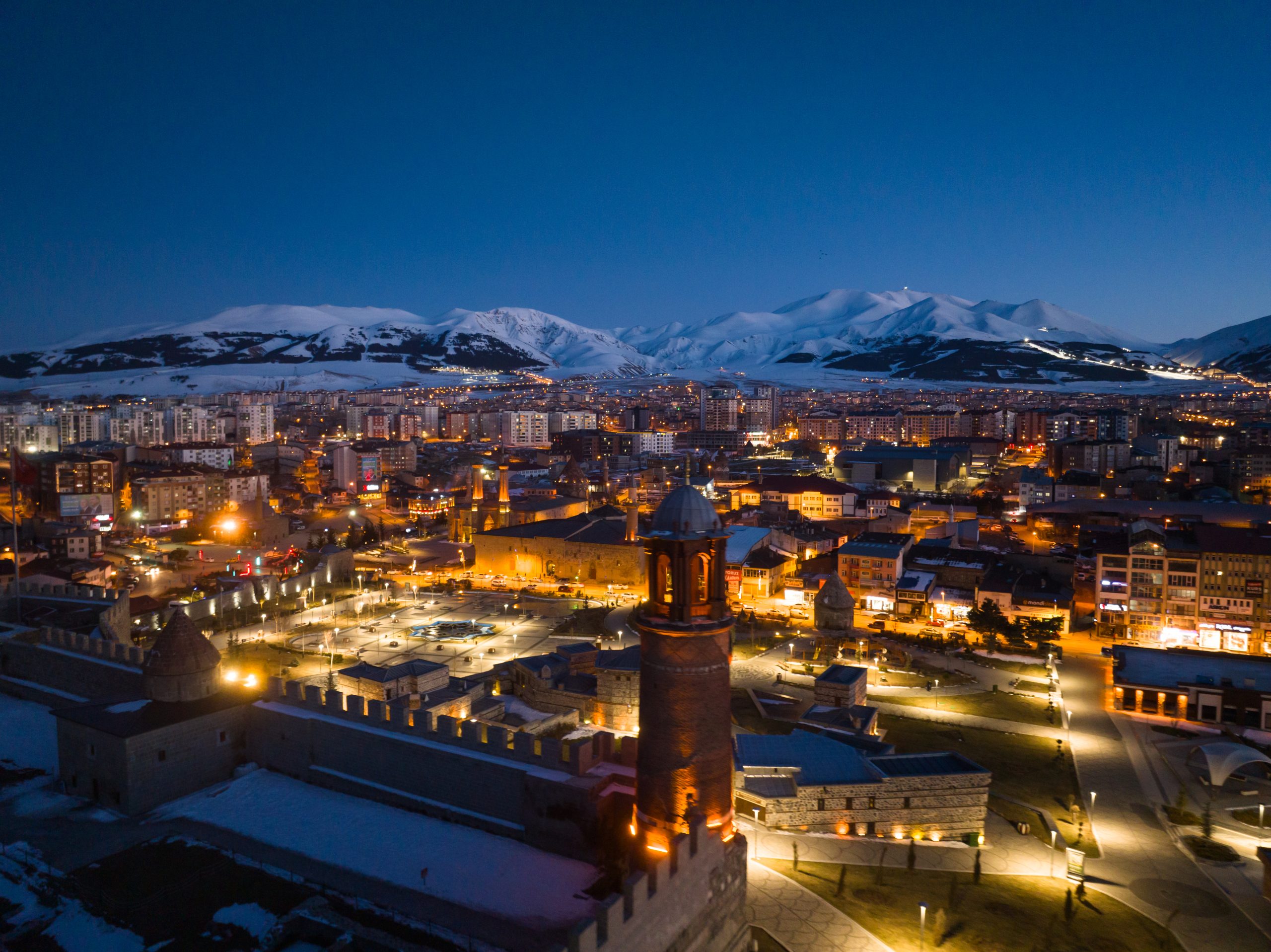Nighttime cityscape with snow-covered mountains in the distance and warm streetlights below.