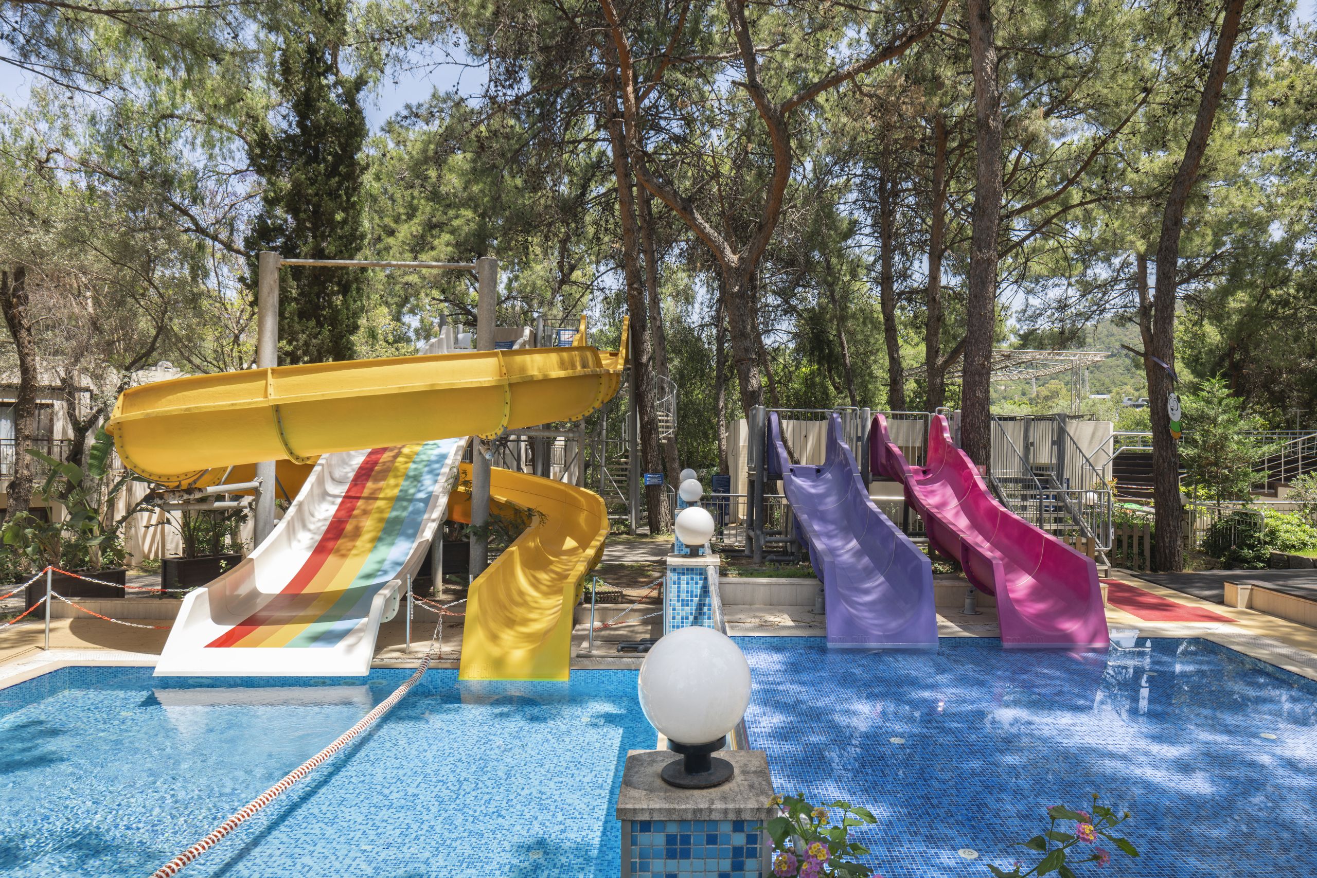Colorful water slides above a blue tiled pool at an outdoor water park, with trees in the background.