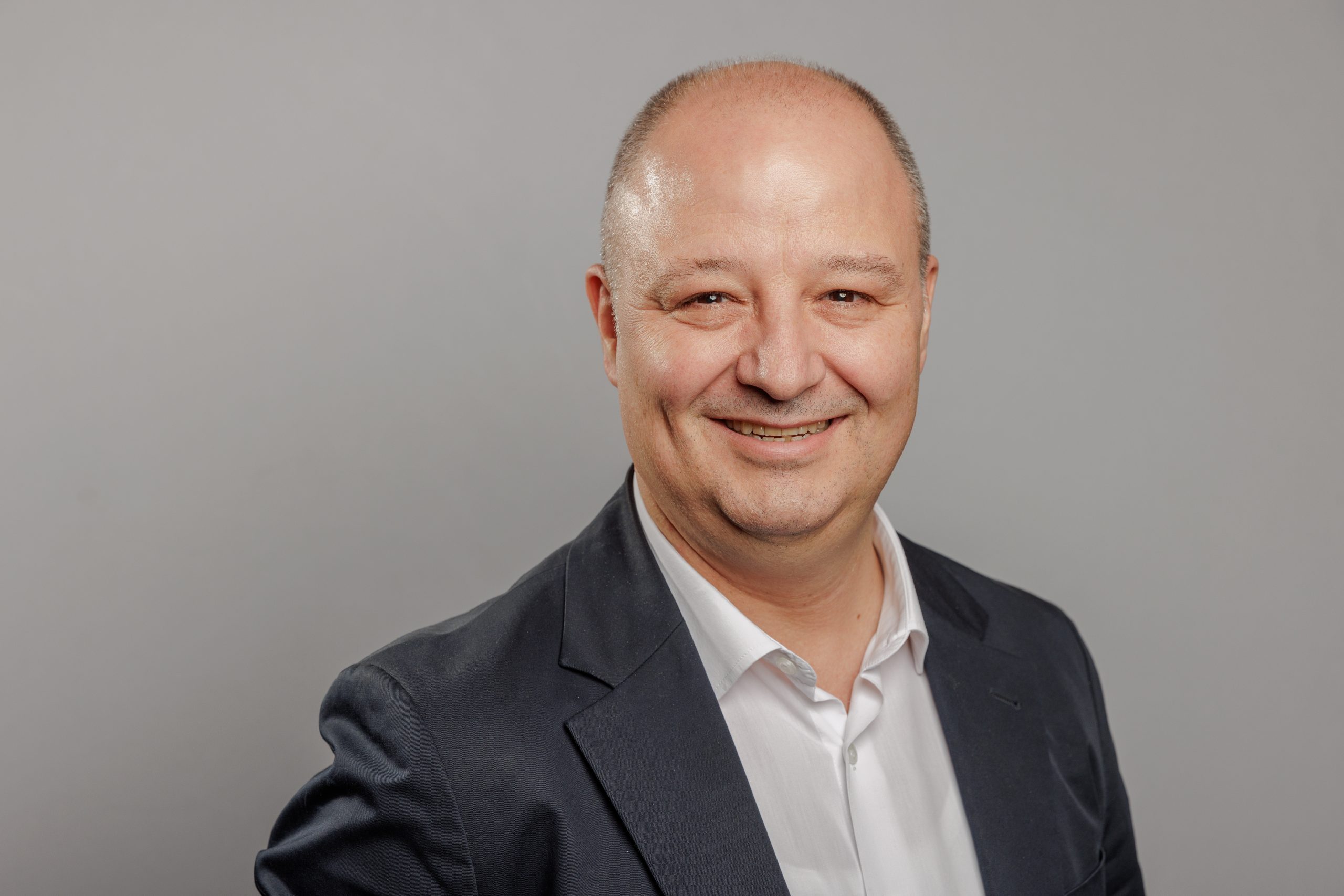 1786515795_GM_Tolga_Aytan | HotelRestaurant Haber Dergisi Smiling man in a dark suit and white shirt posing for a portrait against a gray background.