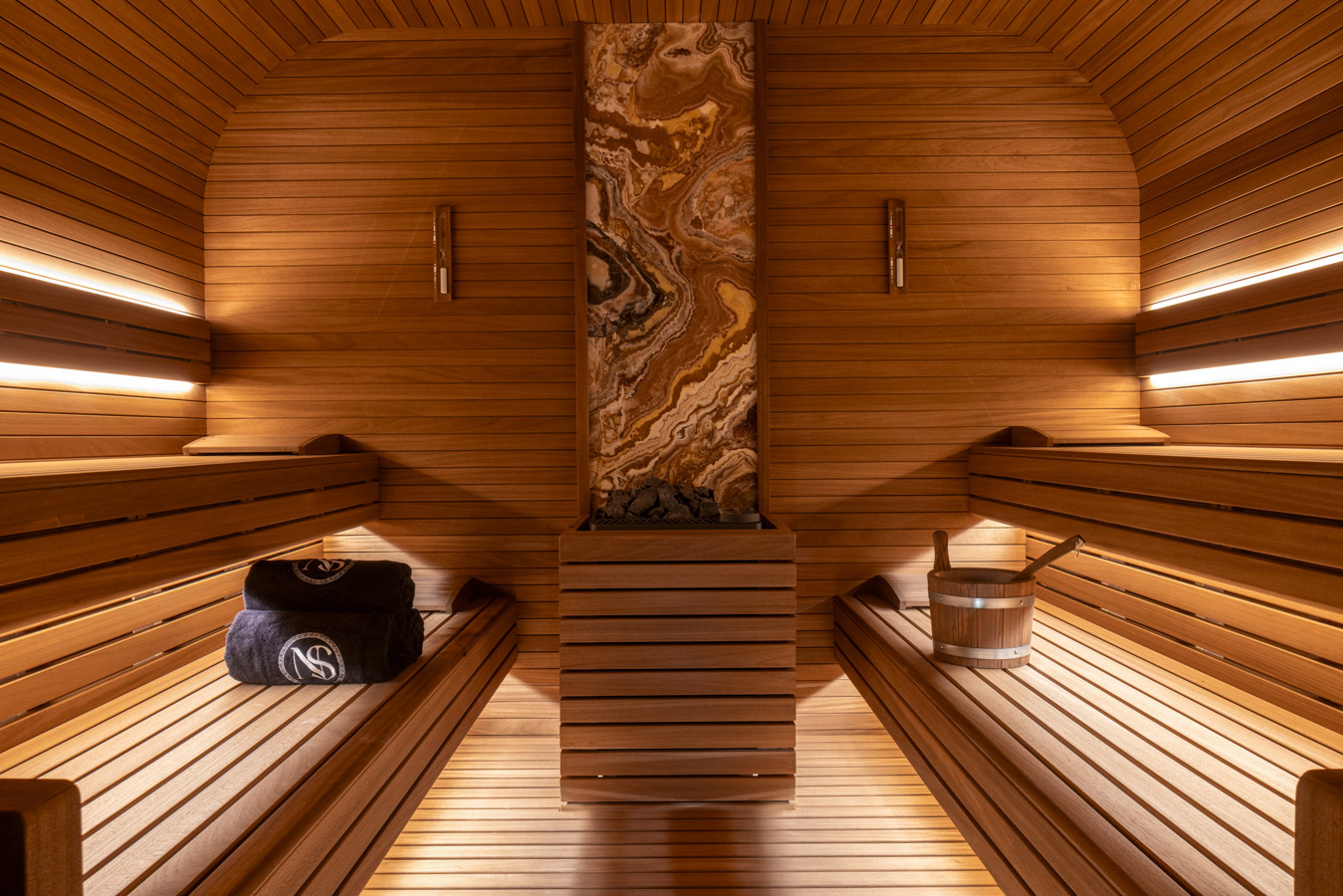 Wood-paneled sauna with a central heater and marble stone surround, warm lighting, towels folded on the bench, and a wooden bucket nearby.