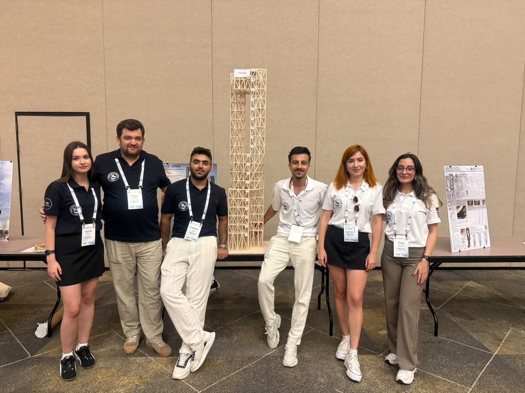 Six students wearing name badges pose beside a tall wooden lattice tower model on a table at an indoor expo.