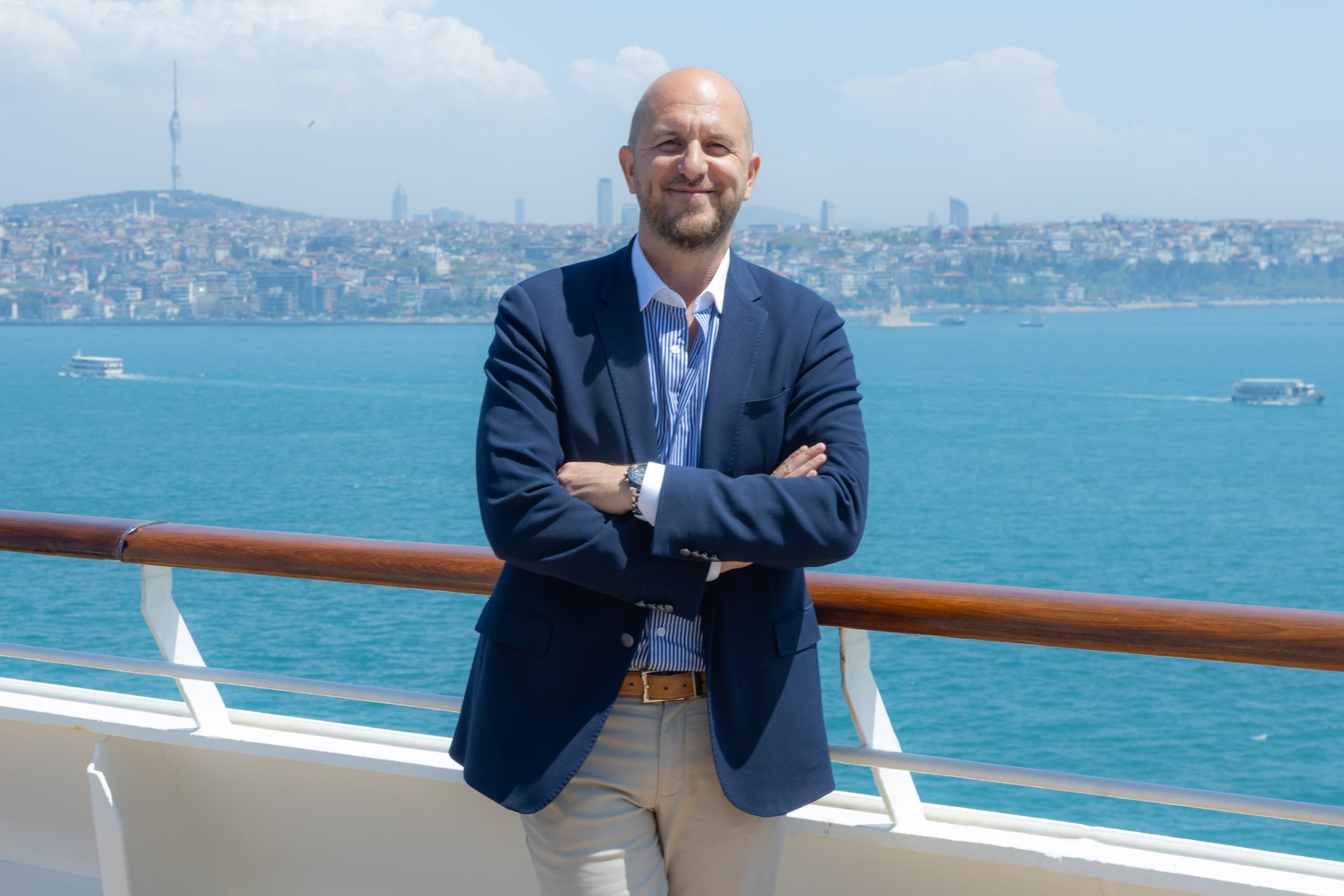 Man in a navy blazer standing with arms crossed on a ship railing, with city and sea in the background.