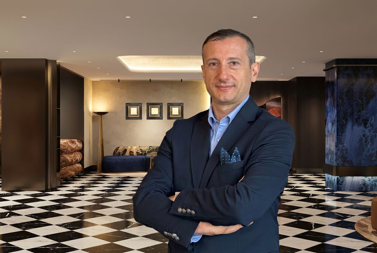 Man in a navy suit with a blue shirt and pocket square stands with arms crossed in a stylish lobby with a black-and-white checkered floor.”] ,