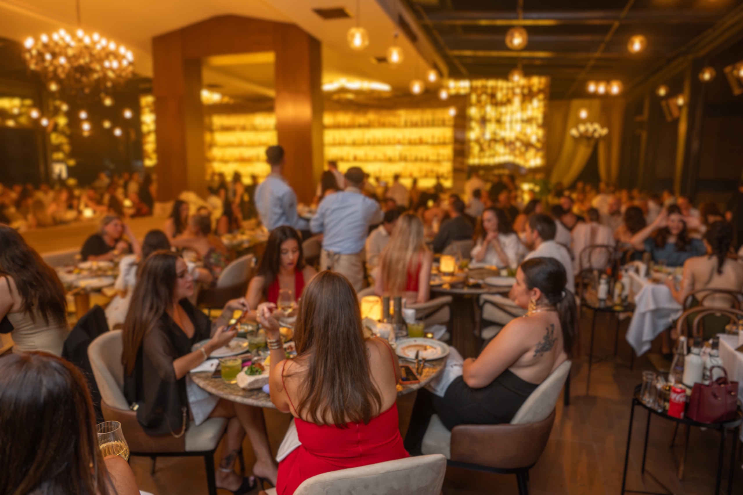 Busy upscale restaurant filled with groups of people dining at round tables under warm, ambient lighting.